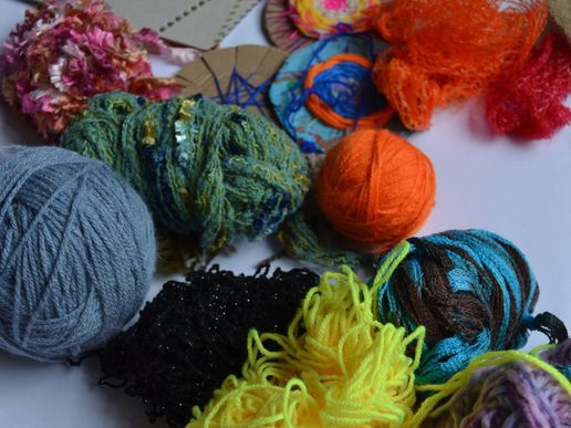 Assorted colourful yarns and textured craft materials spread across a table, including balls of grey and orange wool, bright yellow strands, green and multicoloured fibres, and handmade woven pieces with netting and thread.