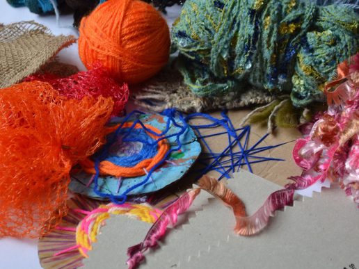 Close-up of colourful craft materials spread across a surface, including bright orange netting, balls of yarn, tangled blue threads, and textured woven fabric pieces in green and pink tones.