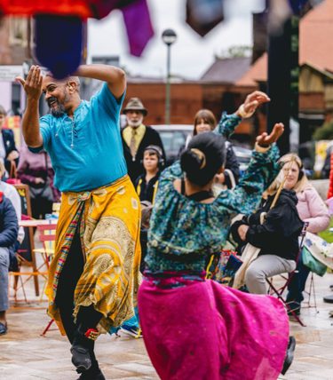 Two performers in colourful costumes dance in an outdoor public space while seated audience members watch from around small café tables during a street arts festival.