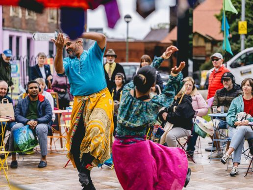 Two performers in colourful costumes dance in an outdoor public space while seated audience members watch from around small café tables during a street arts festival.