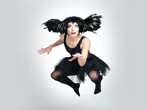 A performer from An Evening Without Kate Bush leaps through the air in a black tutu and dramatic feathered headdress, reaching towards the camera against a white background.