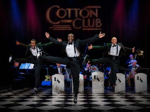 Three male dancers in black tuxedos leap mid-air across a stage during a performance of The Cotton Club. Their arms are outstretched and legs kicked high in a synchronised jazz routine, while a live swing band performs behind them. Colourful red and blue spotlights shine above a black-and-white chequered floor, with the glowing “Cotton Club” logo projected onto the backdrop.