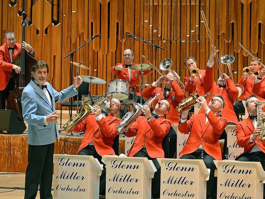 Promotional image for The World Famous Glenn Miller Orchestra directed by Ray McVay, featuring the full big band in bright red jackets playing brass and wind instruments. Centre stage, a female vocalist and drummer are mid-performance, with additional featured performers in tuxedos. The backdrop has a vintage-style banner with blue rays and bold black text.