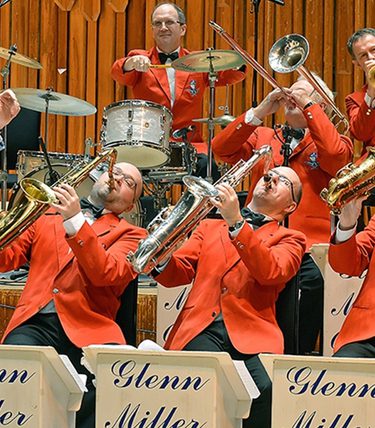 Promotional image for The World Famous Glenn Miller Orchestra directed by Ray McVay, featuring the full big band in bright red jackets playing brass and wind instruments. Centre stage, a female vocalist and drummer are mid-performance, with additional featured performers in tuxedos. The backdrop has a vintage-style banner with blue rays and bold black text.