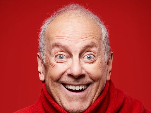 Close-up of Gyles Brandreth, smiling widely against a bright red background, wearing a red turtleneck sweater.