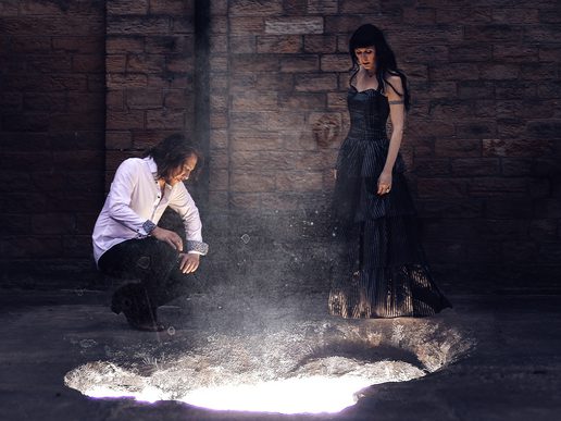 Folk duo Hannah Sanders and Ben Savage in a dramatic setting—Ben crouches beside a glowing crater in the ground, while Hannah stands beside him in a long, dark gown. The scene is set against a textured brick wall, evoking mystery and intrigue. Promotional image for their musical collaboration.