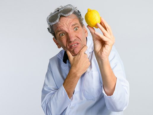 A man in a white lab coat and safety goggles holds a lemon up in one hand while stroking his chin with the other, pulling a confused and exaggerated expression against a plain light background.