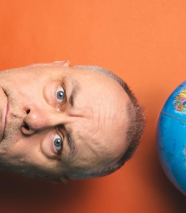 Comedian Jack Dee in a suit and patterned tie leans sideways with his head resting on a flat surface beside a colourful world globe, set against a vibrant orange background.
