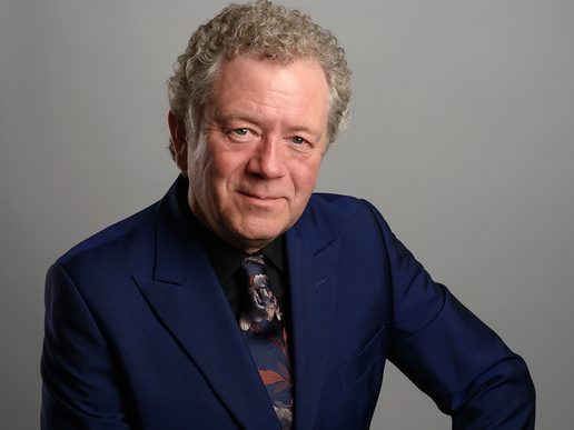 Portrait of comedian and impressionist Jon Culshaw wearing a dark blue suit and patterned tie, smiling slightly against a plain grey background.