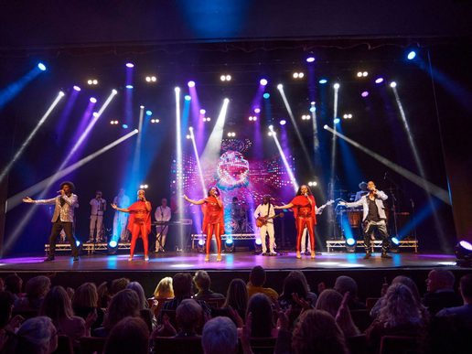 Stage performance of Lost in Music featuring a group of singers and musicians in bright costumes, performing under colourful concert lighting with a disco-style backdrop, as an audience watches from the theatre seats.