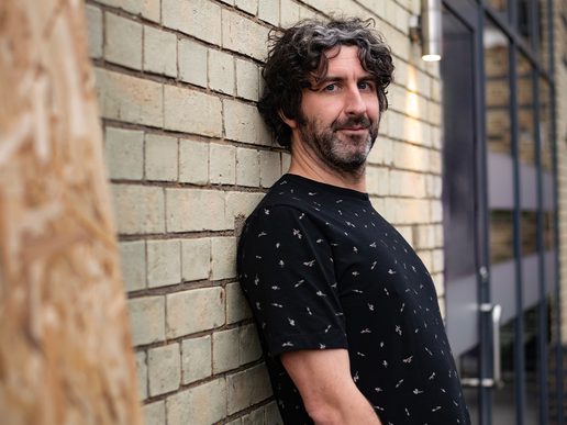 Comedian Mark Watson leans casually against a light brick wall, wearing a black T-shirt patterned with small white symbols. He looks directly at the camera with a relaxed expression, his curly dark hair streaked with grey and a short beard framing his face. A modern glass door and exterior light fixture appear in the background, suggesting an urban setting.