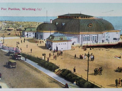 Historic postcard illustration of the Pier Pavilion in Worthing, showing the domed seaside building on the beach with people strolling, early motor cars, and the sea in the background.