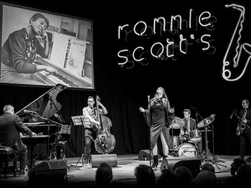 A black-and-white photo of a live jazz performance on stage, featuring a pianist, double bassist, drummer, saxophonist and a female vocalist at the centre. Behind them is a large projected image of Ella Fitzgerald at a piano, and the illuminated “Ronnie Scott’s” jazz club logo glows on the right.