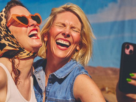 Scummy Mummies laugh together in the sunshine, one wearing heart-shaped sunglasses and a patterned headscarf, as they pose for a selfie.