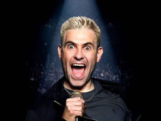 Simon Brodkin stands under a spotlight on a dark stage, holding a microphone and shouting with a wide, animated expression, eyes open wide and mouth open mid-performance.