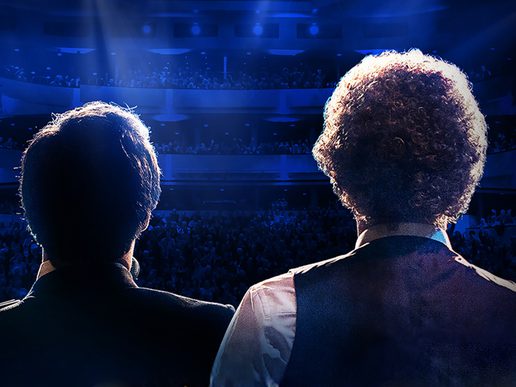 View from behind two performers standing on stage under blue spotlights, facing a packed theatre audience. One has short dark hair, and the other has curly light hair, evoking the iconic duo Simon & Garfunkel. The atmosphere is dramatic and celebratory, capturing a live concert moment.