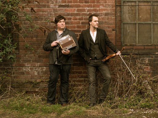 Spiers & Boden stand against a weathered brick wall, one holding a button accordion and the other a fiddle and bow, surrounded by overgrown plants.