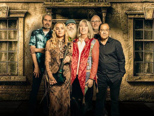 Six members of folk rock band Steeleye Span stand together in front of a rustic cottage backdrop. A young woman at the front holds a violin, while the others—four men and one woman—stand close behind, dressed in a mix of patterned shirts and colourful jackets, facing the camera with calm, confident expressions.