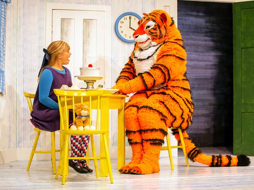 A girl with blonde hair tied in a ribbon sits at a small yellow table, talking to a large friendly tiger in an orange and black costume seated opposite her. A toy tiger sits on the chair beside her. The scene takes place in a colourful, childlike kitchen set.