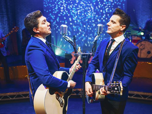 Two performers dressed in blue suits play guitars and sing into vintage microphones on stage, recreating the style of The Everly Brothers in Walk Right Back – The Everly Brothers Story.