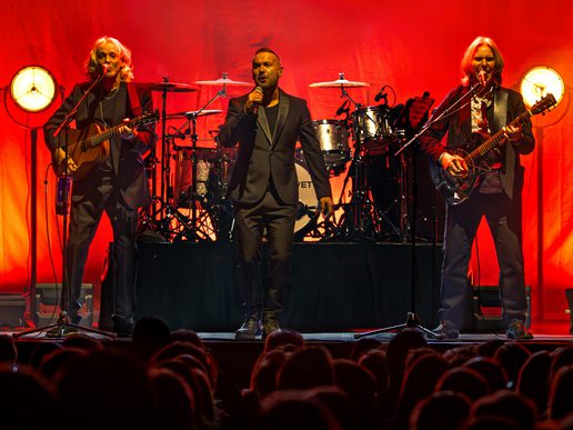 Wet Wet Wet perform live on stage against a dramatic red backdrop. The lead singer stands center-stage holding a microphone, flanked by two guitarists, each playing under warm vintage-style lights. A drum kit is visible behind them, and the silhouettes of a captivated audience fill the foreground.