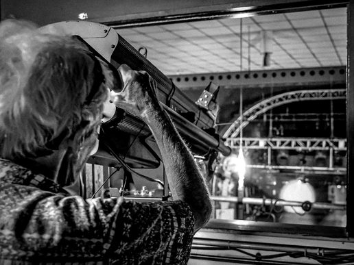 A man on a backstage theatre tour looks through a large stage spotlight from a control booth overlooking an empty auditorium. The black-and-white photograph captures the behind-the-scenes perspective, with lighting trusses and stage equipment visible through a large viewing window.