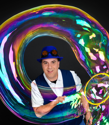 A colourful bubble artist dressed in a blue hat, goggles, and matching vest creates a giant, iridescent soap bubble that loops dramatically around him, set against a black background.