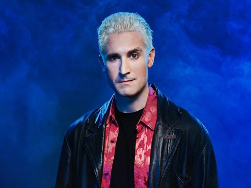 Portrait of a pale-skinned man with short, platinum blond hair, wearing a black leather jacket over a red patterned shirt. He faces forward with a serious expression against a smoky blue background.