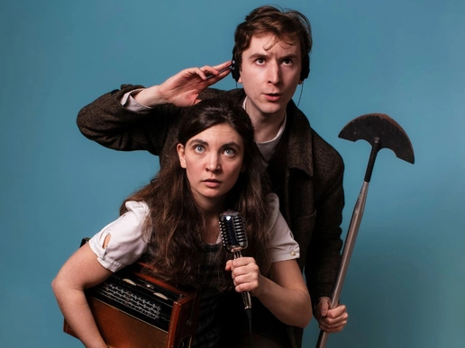Two performers from Down to Chance pose against a blue background, with one holding a vintage microphone and radio while the other listens through headphones and holds a long-handled prop tool, both looking alert and curious.