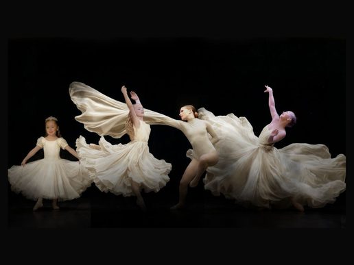 Four dancers in flowing cream costumes perform on a dark stage, captured mid-movement with sweeping skirts and expressive poses, creating a sense of motion and elegance.