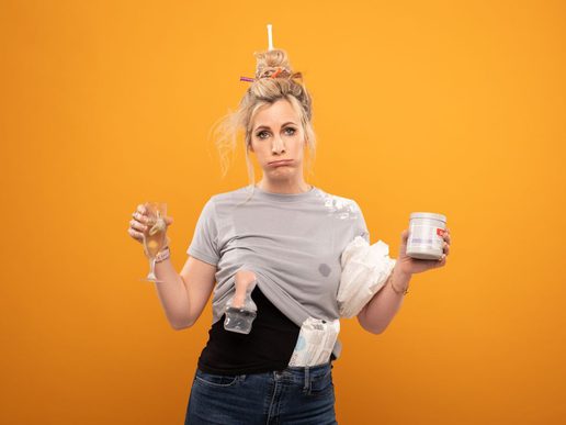Performer Hannah East stands against a bright orange background, looking exasperated while holding a glass of wine and a container, with baby-related items tucked into her clothing and hair in a messy bun.