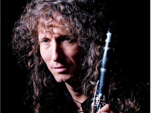 Portrait of clarinettist Julian Marc Stringle with long curly hair, holding a clarinet against a dark background and looking thoughtfully towards the camera.