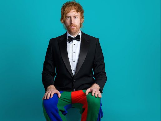 A man with tousled ginger hair and light stubble sits facing forward against a plain turquoise background. He wears a black tuxedo jacket, white dress shirt, and black bow tie, paired with brightly coloured trousers in red, green, and blue panels. His hands rest on his knees and he looks directly at the camera with a neutral, slightly serious expression.