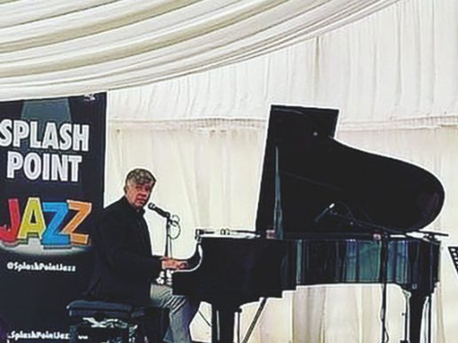 Pianist Neal Richardson performing at a grand piano with a microphone, beside a “Splash Point Jazz” banner inside a white marquee venue.
