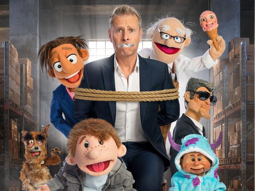 Ventriloquist Paul Zerdin stands at the centre of a warehouse surrounded by his colourful puppet characters. Wearing a dark suit, he has tape over his mouth and is tied around the waist with thick rope, looking wide-eyed and bewildered. Around him are a mischievous puppet in a blue jacket, an elderly scientist holding an ice cream topped with a baby puppet's head, a sharp-suited puppet wearing sunglasses, a scruffy dog puppet, a cheerful young boy puppet and a smiling baby dressed in a blue monster costume. Tall warehouse shelving stacked with boxes fills the background, creating a playful scene that suggests chaos and comedy.