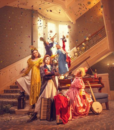 A lively promotional image for Pride and Prejudice featuring six performers in colourful Regency-inspired costumes arranged across a grand staircase and elegant drawing room. Confetti rains through the air as musicians play on the staircase, while one performer stands confidently at the front holding a red book. Around them are a piano, accordion, banjo, lanterns and discarded clothing, hinting at a fast-paced, music-filled adaptation. Warm golden lighting, ornate architecture and theatrical poses create a joyful, energetic atmosphere blending period drama with playful comedy.