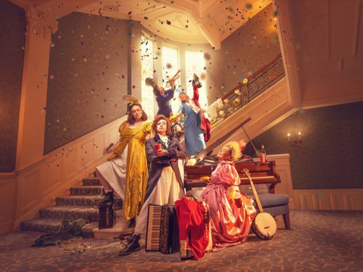 A lively promotional image for Pride and Prejudice featuring six performers in colourful Regency-inspired costumes arranged across a grand staircase and elegant drawing room. Confetti rains through the air as musicians play on the staircase, while one performer stands confidently at the front holding a red book. Around them are a piano, accordion, banjo, lanterns and discarded clothing, hinting at a fast-paced, music-filled adaptation. Warm golden lighting, ornate architecture and theatrical poses create a joyful, energetic atmosphere blending period drama with playful comedy.