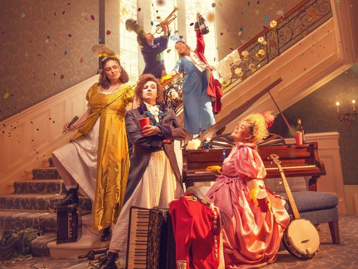 A lively promotional image for Pride and Prejudice featuring six performers in colourful Regency-inspired costumes arranged across a grand staircase and elegant drawing room. Confetti rains through the air as musicians play on the staircase, while one performer stands confidently at the front holding a red book. Around them are a piano, accordion, banjo, lanterns and discarded clothing, hinting at a fast-paced, music-filled adaptation. Warm golden lighting, ornate architecture and theatrical poses create a joyful, energetic atmosphere blending period drama with playful comedy.