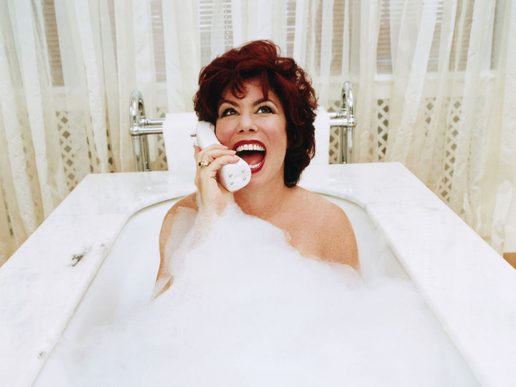 Comedian Ruby Wax sits in a bubble-filled bath, smiling widely while holding a phone to her ear, with white curtains and a bright bathroom setting behind her.