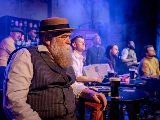 A group of performers sit around small pub tables with pints of stout, creating the atmosphere of a traditional Irish pub. In the foreground, a bearded man in a waistcoat and flat-brimmed hat looks thoughtfully towards the stage, while the others watch on from behind. Warm lighting, blue stage haze and authentic pub décor evoke the lively setting of Seven Drunken Nights – The Story of The Dubliners.