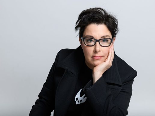 Portrait of Sue Perkins wearing a black jacket and glasses, resting her head on her hand and looking directly at the camera against a plain light background.