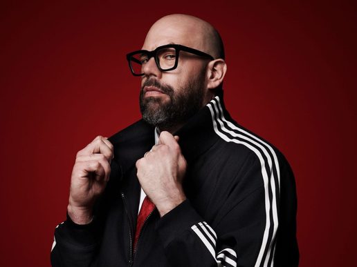 Comedian Tom Davis poses against a deep red background, wearing black-framed glasses and a black tracksuit jacket with white stripes.