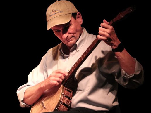 A man wearing a baseball cap and light button-down shirt plays a banjo on stage, looking down in concentration. Warm stage lighting highlights his face and hands against a dark background.
