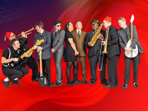 image for Too Much Too Young featuring nine ska musicians standing and crouching in a line against a vivid red background. The group wear smart black and grey suits, hats and sunglasses, with several holding instruments including electric guitars, trumpet, saxophone and trombone. One musician wears a bright red fez hat while another holds a white guitar, creating a playful, energetic band portrait inspired by classic ska style.