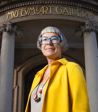 Incoming Worthing Theatres & Museum CEO Louise Blackwell stands outside Worthing Museum & Art Gallery, wearing a bright yellow coat and blue glasses. The museum’s grand stone entrance, columns and gold lettering are visible behind her.