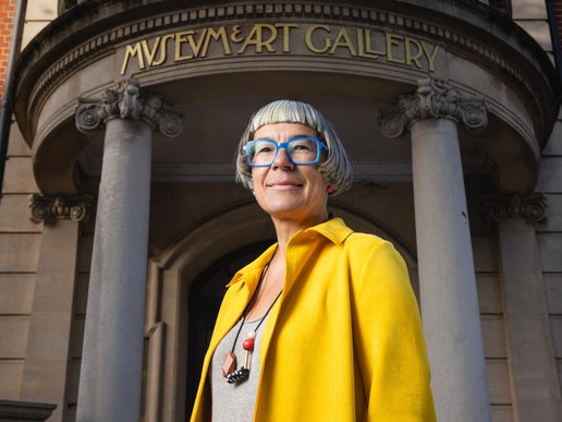 Incoming Worthing Theatres & Museum CEO Louise Blackwell stands outside Worthing Museum & Art Gallery, wearing a bright yellow coat and blue glasses. The museum’s grand stone entrance, columns and gold lettering are visible behind her.