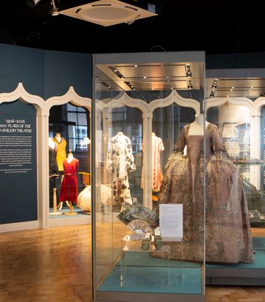 Wide view of the Historic Dress gallery at Worthing Museum, featuring elegant period garments displayed in illuminated glass cases. A richly embroidered 18th-century-style gown takes centre stage, surrounded by historic dresses, accessories and textiles from different eras. Decorative archways frame the exhibition, while interpretive panels explain the history of dressmaking, Worthing's textile heritage, and the Pavilion Theatre's centenary. The spacious gallery has polished wooden flooring, soft lighting, and a calm, contemporary museum setting.