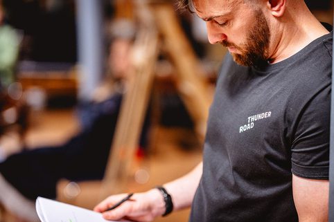 Alex Moran in rehearsal, wearing a black "Thunder Road" t-shirt, looks down and studies a printed script with a pen in hand. The background is softly blurred, suggesting a creative or studio environment.