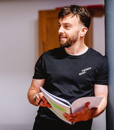 Alex Moran stands casually in a rehearsal space, leaning against a pillar while holding a marked-up script and pen. He is wearing a black "Thunder Road" t-shirt and looking to the side with a relaxed, focused expression.