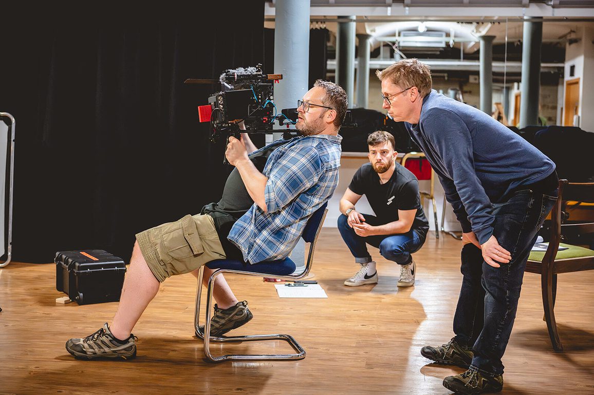 Three men in a rehearsal or filming space focus on a camera setup. One man sits on a chair operating a large professional camera, while another leans in to observe. In the background, Alex Moran crouches attentively, watching the action unfold. Equipment cases and rehearsal materials are scattered on the floor.